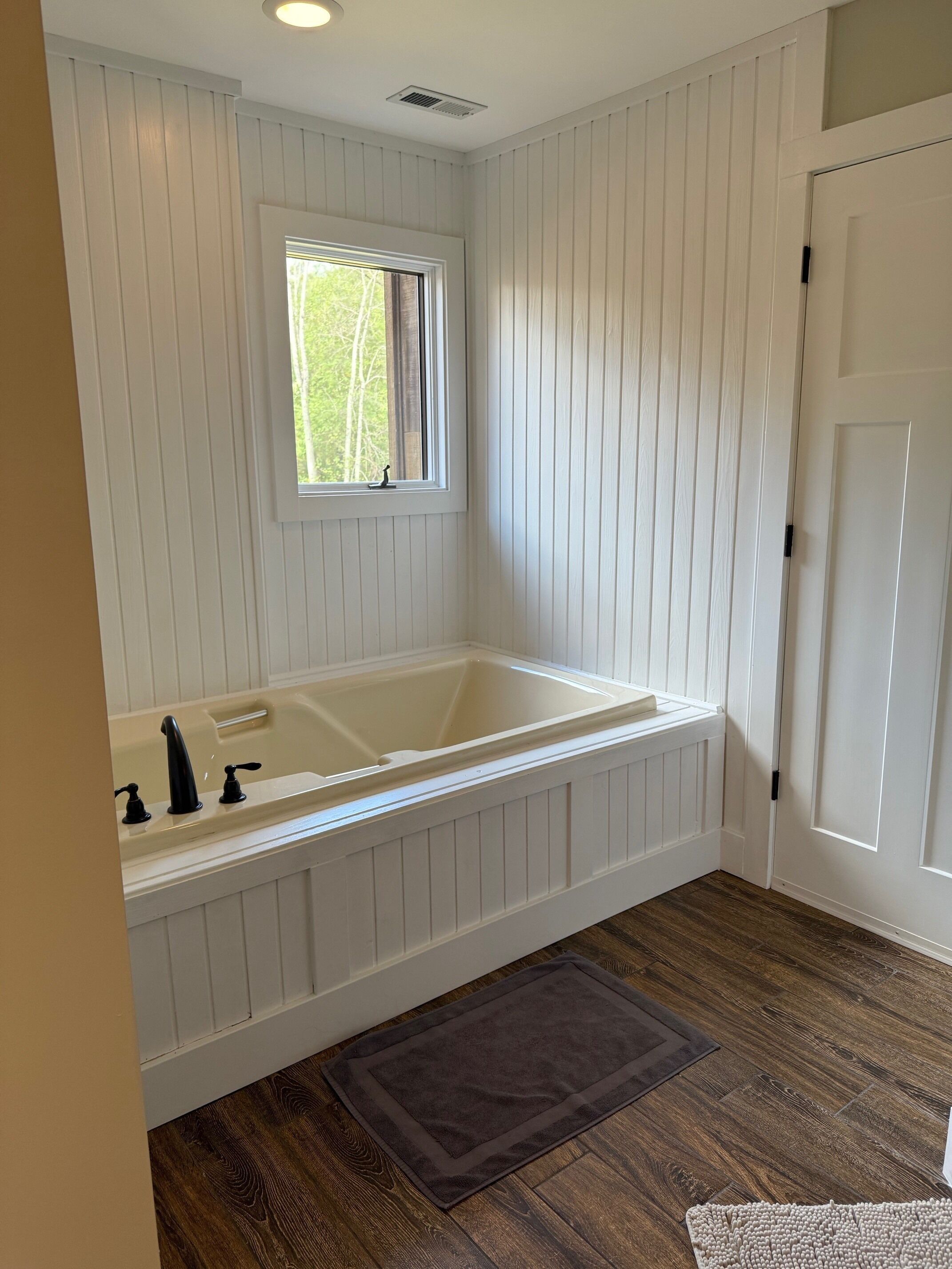 A bathroom with a white bathtub, a window, and wooden flooring.