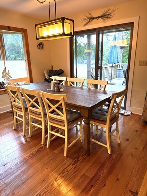 Uma sala de jantar com mesa e cadeiras de madeira, luminária pendente e vista para um deck externo através de amplas janelas.