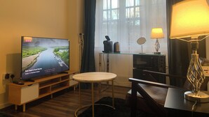 Habitación de hotel con televisión mostrando un programa, una mesita redonda pequeña, una lámpara, un espejo y una ventana con cortinas.