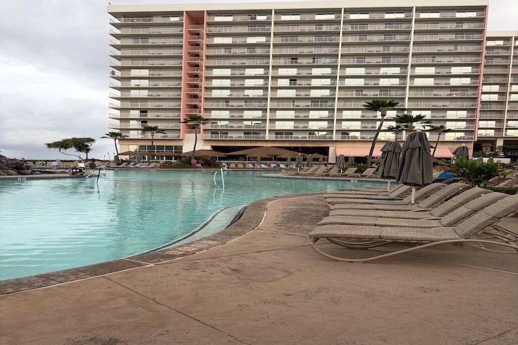 Une piscine d’hôtel avec des chaises longues et des parasols, et un grand bâtiment avec des balcons en arrière-plan.