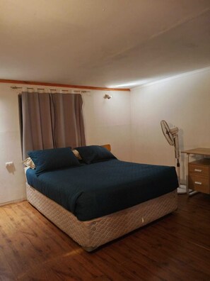 Ein Einzelzimmer mit Holzboden, einem Schreibtisch mit Schubladen, einem Standventilator und einem Fenster mit Vorhängen.