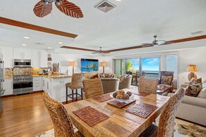 A spacious kitchen with modern appliances, a dining area with wicker chairs, and a view of the ocean through large windows. - Oceanfront Luxury at Lanikai 132 | Central A/C & King Suites (Kapaa)