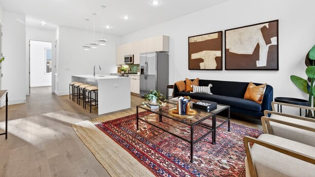 Una sala moderna con sofá, mesa de centro y sillas. Se aprecia una cocina con armarios y electrodomésticos al fondo.