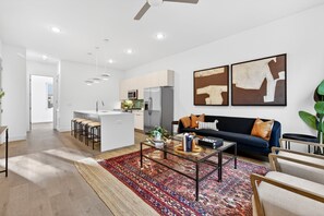 Ein modernes Wohnzimmer mit Sofa, Couchtisch und Sesseln. Im Hintergrund ist eine Küchenzeile mit Schränken und Geräten zu sehen.