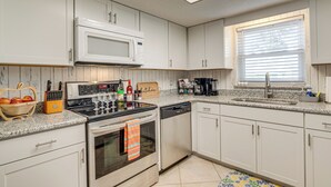 Una cocina moderna con armarios blancos, microondas, estufa y lavavajillas. Hay una ventana con persianas, un fregadero y varios utensilios de cocina sobre la encimera.