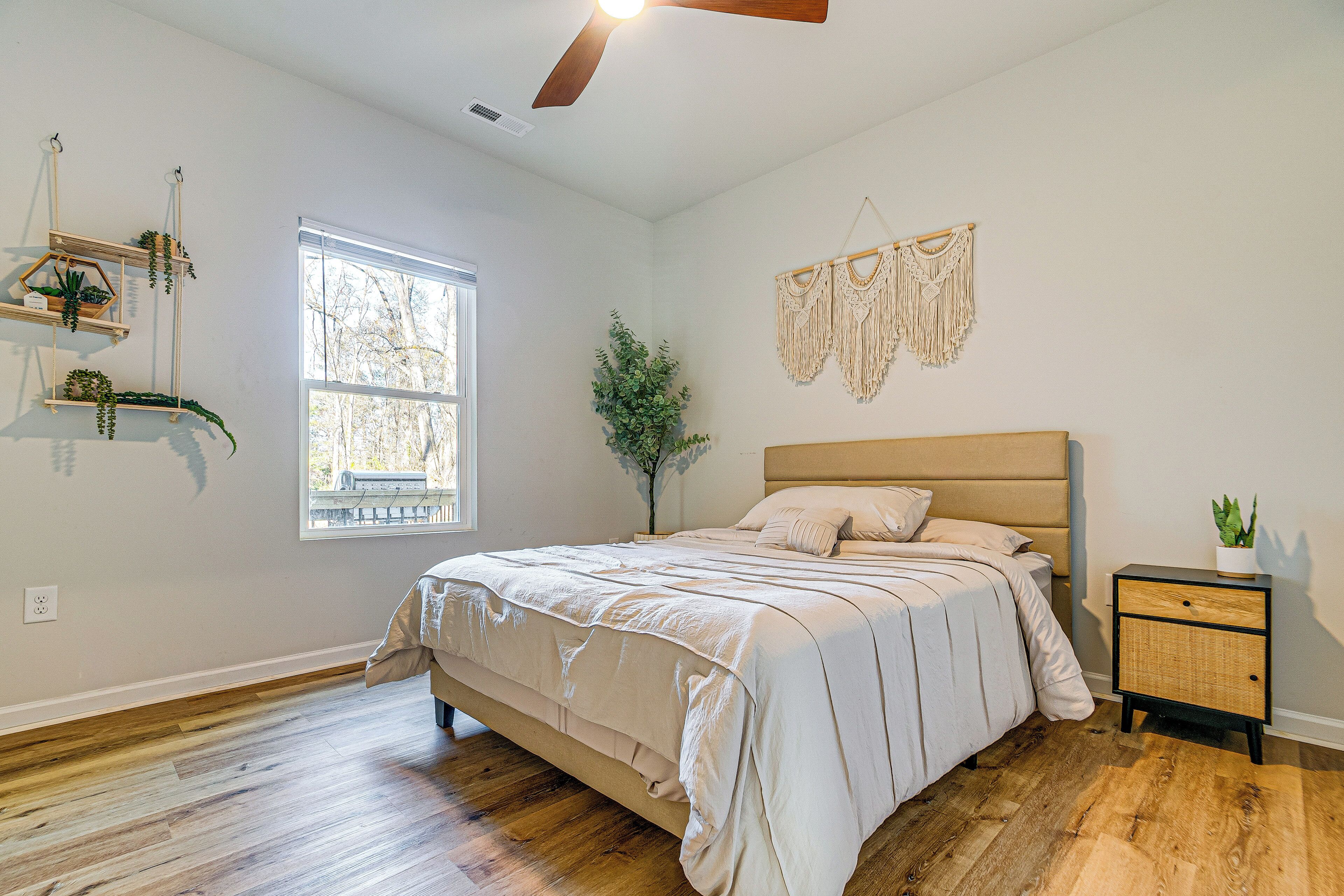 A bedroom with a bed, a nightstand, a wall-mounted macrame piece, and a window with a view of trees.