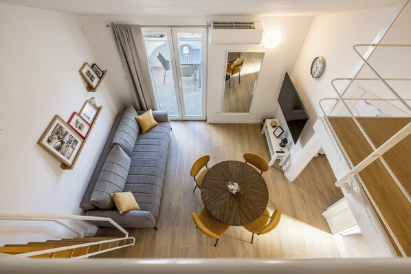 Ein modernes Wohnzimmer mit einem grauen Sofa, einem runden Holztisch mit Stühlen, einem Fernseher und einer Treppe, die zu einer oberen Etage führt.