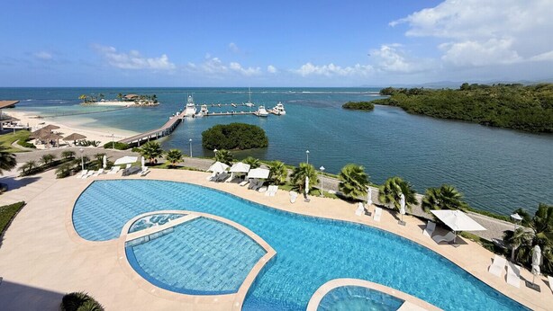 A resort pool with clear blue water, surrounded by lounge chairs and umbrellas, with a view of the sea and boats.