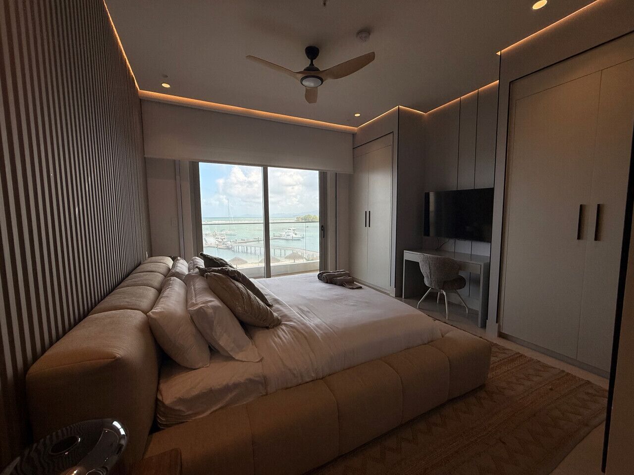 A modern bedroom with a large bed, a seating area, a TV, and a view of the sea.