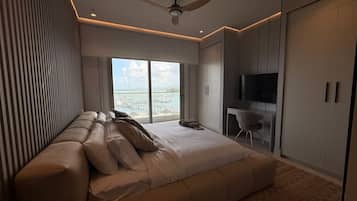 A modern bedroom with a large bed, a seating area, a TV, and a view of the sea.