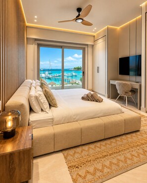 A modern bedroom with a large bed, a desk, a TV, and a view of a marina.