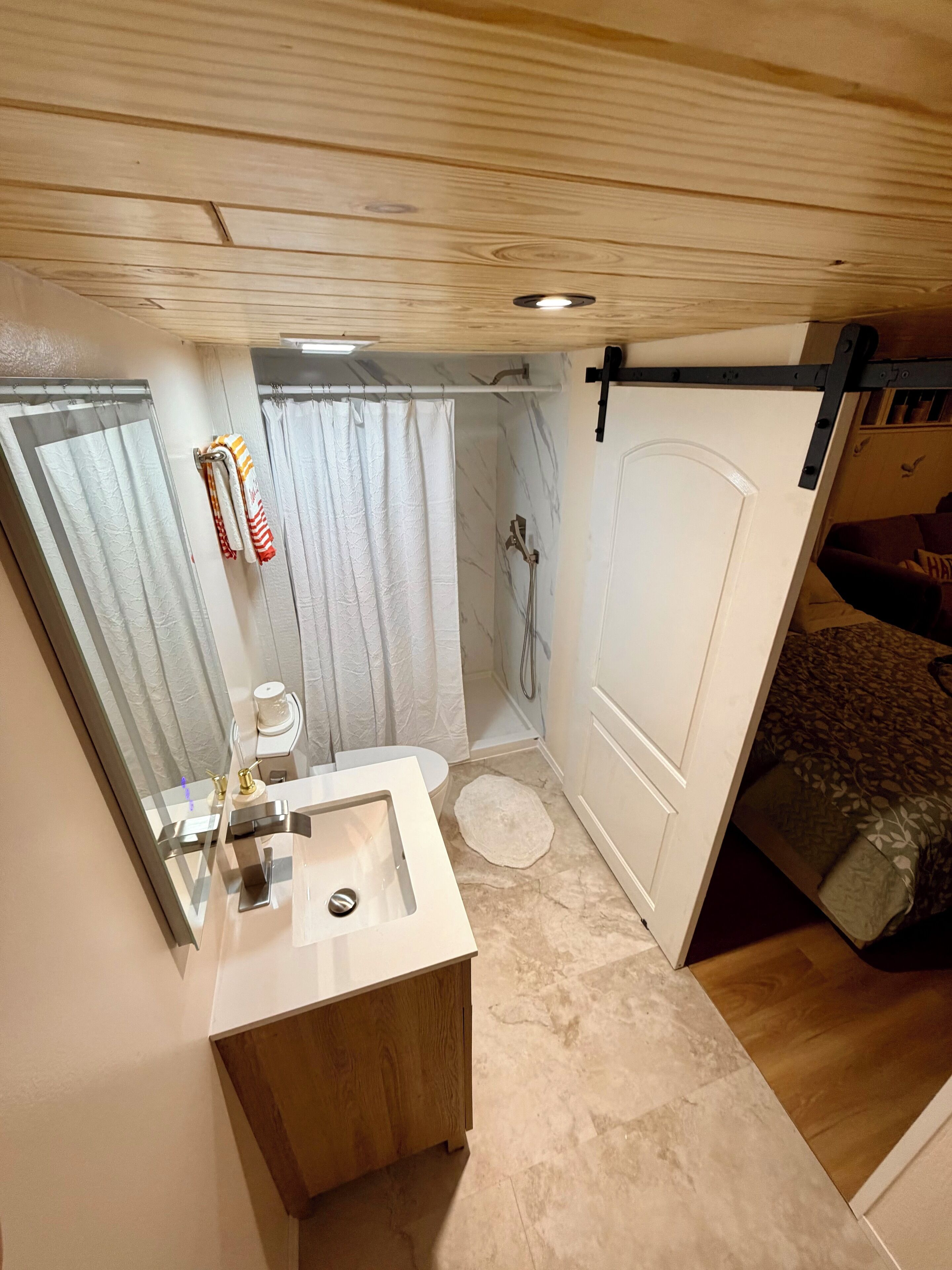 A compact bedroom with a wooden ceiling, a small bathroom with a sink and shower, and a bed with a patterned blanket.