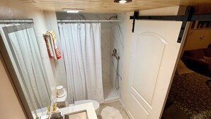 Habitación compacta con techo de madera, baño pequeño con lavamanos y ducha, y cama con manta estampada.