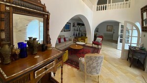 A spacious living area with a high ceiling, a wooden coffee table, a sofa, and a red armchair. There is a large mirror on the left and a staircase leading to an upper level. - Maison Lella Kmar Sidi Bousaid (Site archéologique de Carthage)
