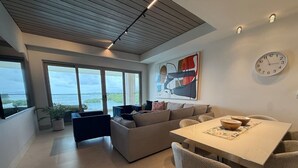 Ein modernes Wohnzimmer mit großem Fenster, einer Couch, einem Esstisch mit Stühlen und einer Wanduhr.