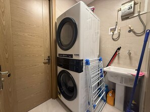 A compact laundry room with a front-loading washer and dryer, a metal drying rack, a sink, and a hose for water connections.
