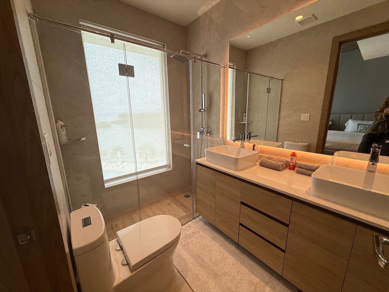 Un baño moderno con cabina de ducha de vidrio, un ventanal grande, tocador doble y un inodoro.