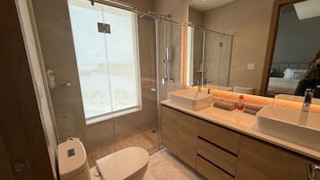 Un baño moderno con cabina de ducha de vidrio, un ventanal grande, tocador doble y un inodoro.