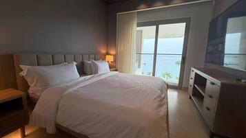 Habitación de hotel con una cama grande, una mesita de noche, un aparador y vistas al océano a través de un ventanal.