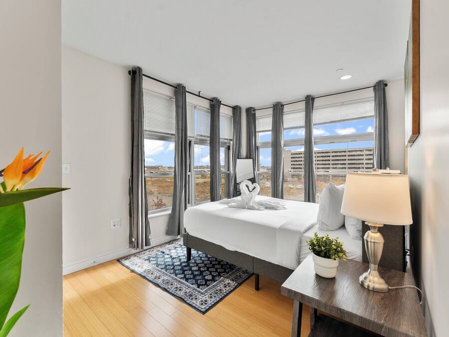 Una habitación de hotel con cama, escritorio, lámpara, planta en maceta y ventanas grandes con persianas.