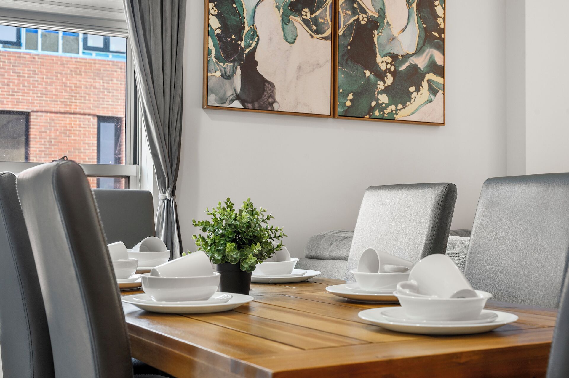 Un coin repas avec une table en bois dressée pour quatre personnes, des chaises à revêtement gris, une petite plante en pot et une œuvre d’art abstraite accrochée au mur.
