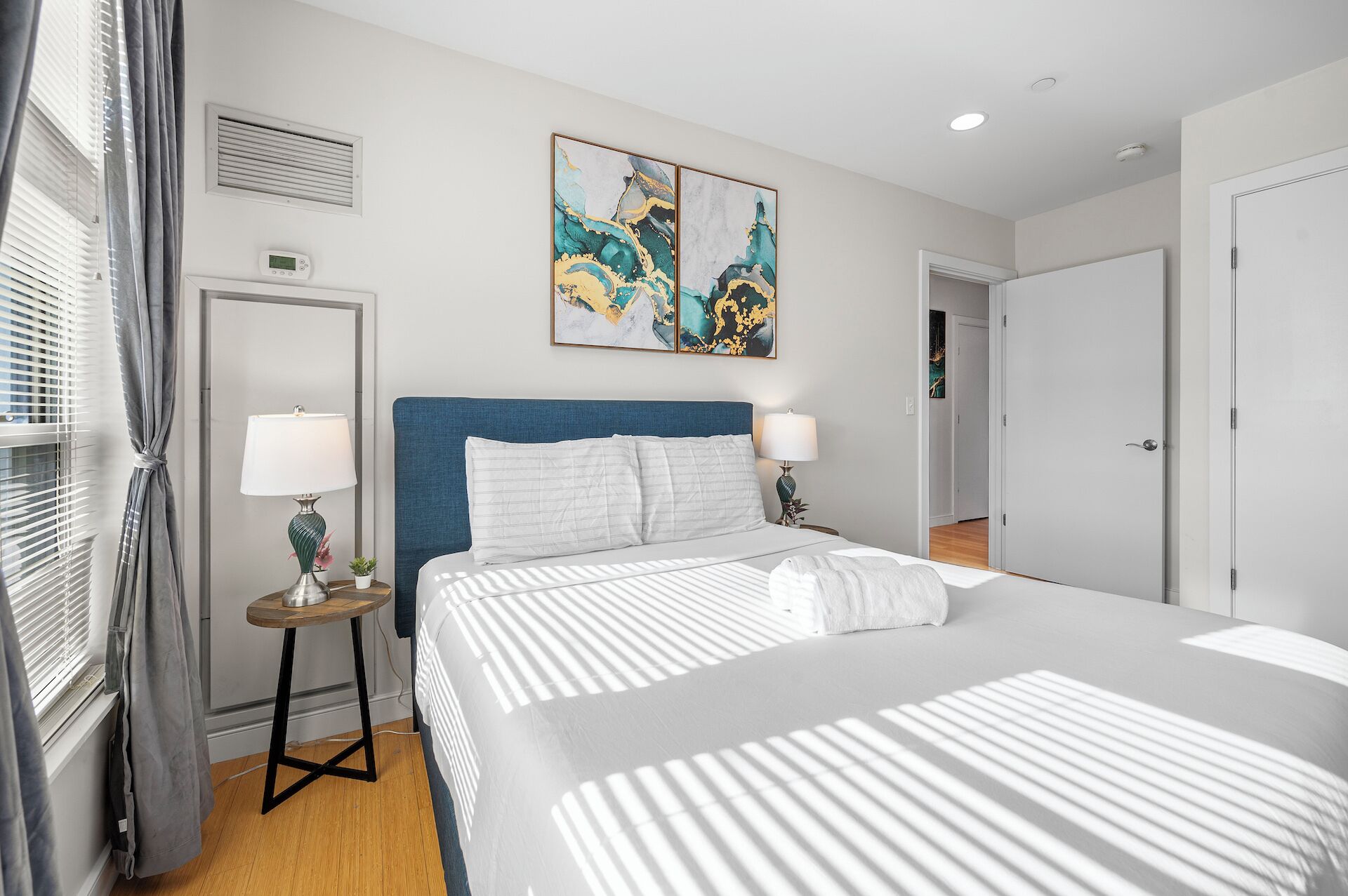 A hotel room with a large bed, two bedside tables with lamps, a window with curtains, a painting on the wall, and a door to another room.