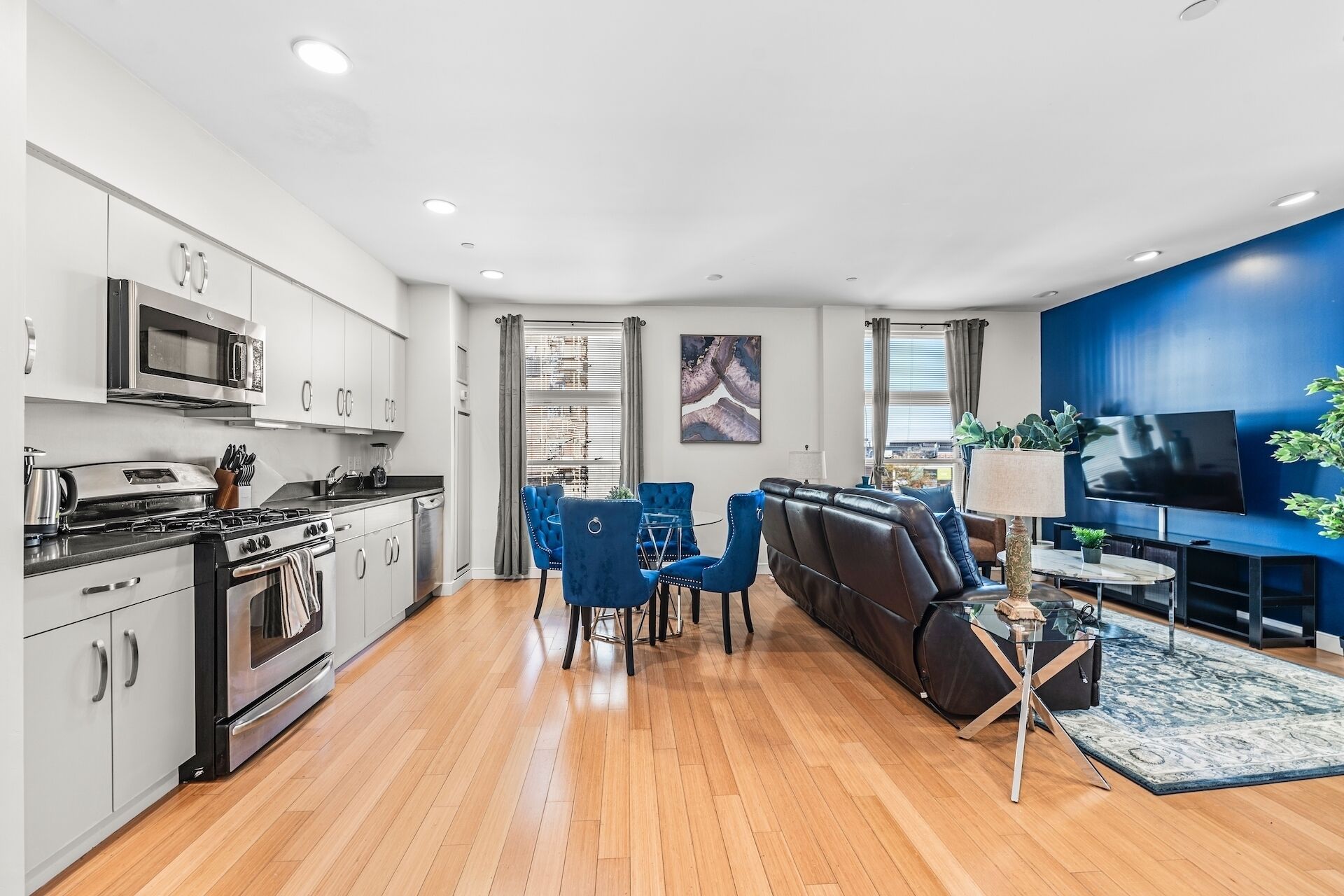 A modern kitchen with stainless steel appliances, a dining area with blue chairs, a leather sofa, and a living room with a TV and plants.