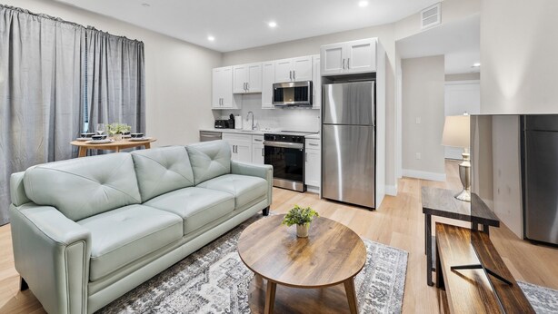 A modern living room with a sofa, a dining table, and a kitchen area with appliances. - Luxury 2BR near Cambridge (Somerville)