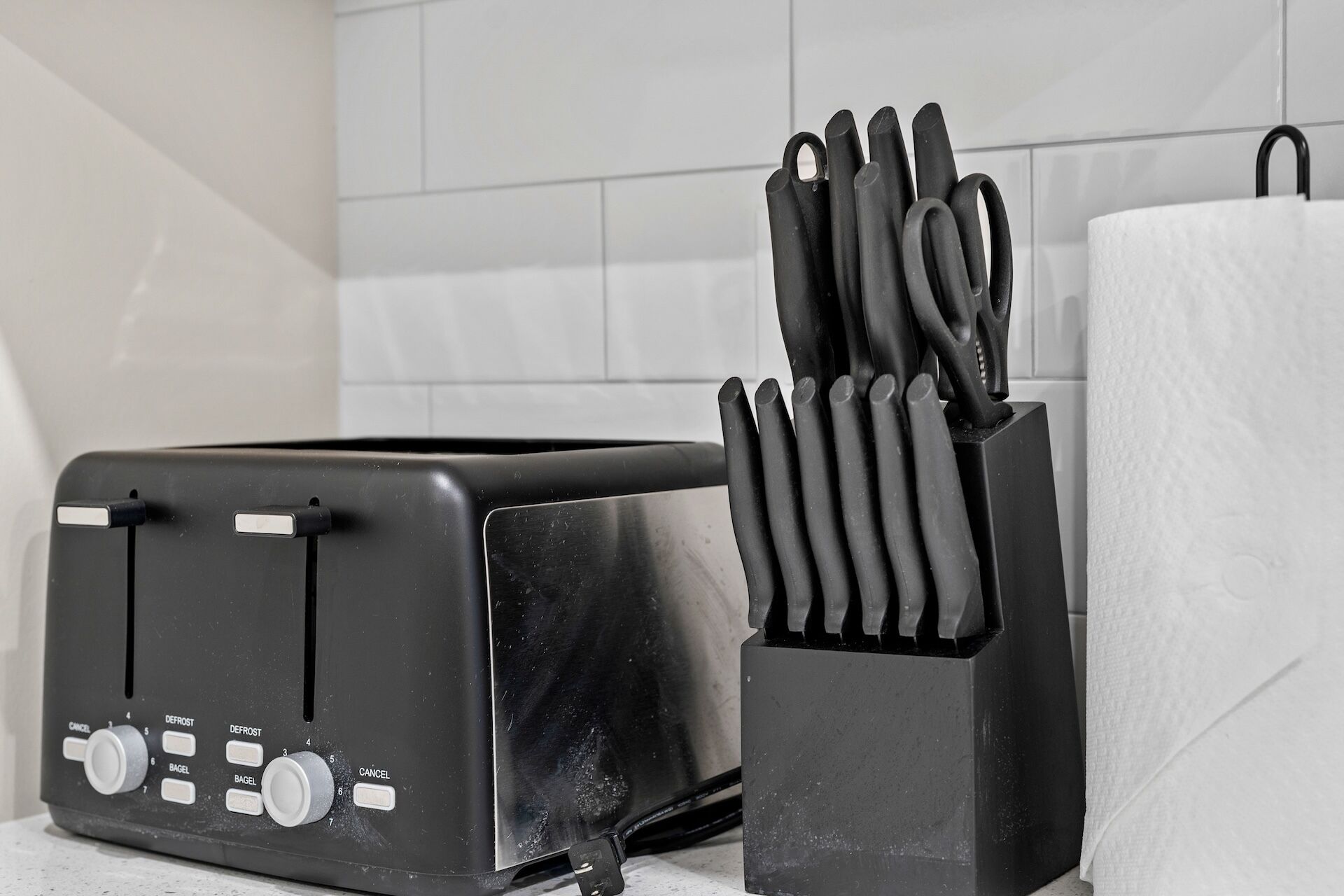 A kitchen with a black toaster, a paper towel holder with utensils, and a white tiled backsplash.