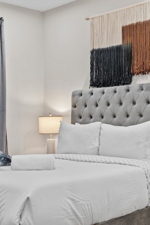 A bed with white bedding, a tufted headboard, and a wall-mounted macrame piece. - Luxury 2BR near Cambridge (Somerville)