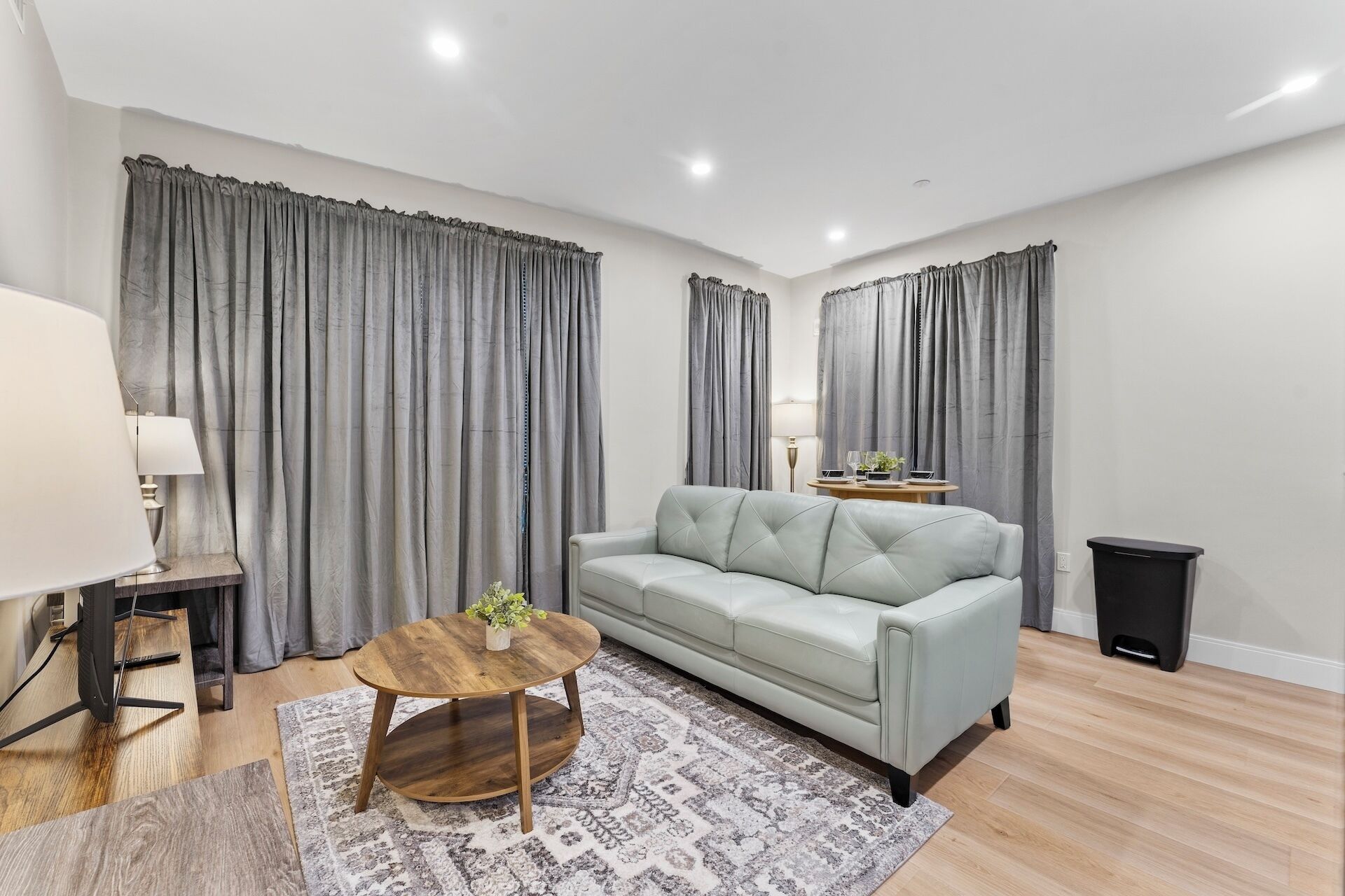 Ein modernes Wohnzimmer mit einem hellgrauen Sofa, einem runden Couchtisch aus Holz und grauen Vorhängen.