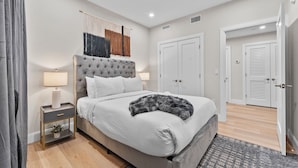 A bedroom with a tufted headboard, a bed with white bedding, a bedside table with a lamp, and a wall-mounted macrame piece. - Luxury 2BR near Cambridge (Somerville)