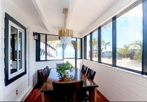 Comedor con una larga mesa de madera, sillas, una araña y ventanas amplias con vista a la playa.