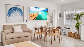 A modern living room with a beige sofa, a dining table with chairs, a large TV, and a window with blinds. - Hawksbill 201 | Beach Studio | Heated Pool (Lauderdale-by-the-Sea)