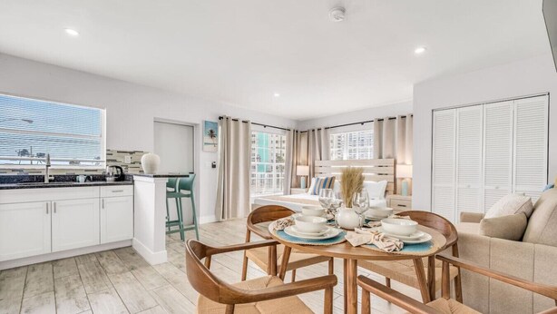 A modern kitchen with white cabinets, a dining area with wooden chairs and a round table set for four, and a bedroom with a large window and a bed with white linens. - Hawksbill 201 | Beach Studio | Heated Pool (Lauderdale-by-the-Sea)