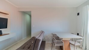 Un salon moderne et minimaliste, comprenant un canapé, une table à manger avec des chaises, un téléviseur à écran plat fixé au mur et une petite étagère en bois.