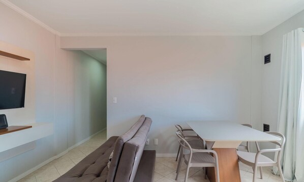 Una sala de estar moderna y minimalista con un sofá, una mesa de comedor con sillas, un televisor de pantalla plana montado en la pared y un pequeño estante de madera.