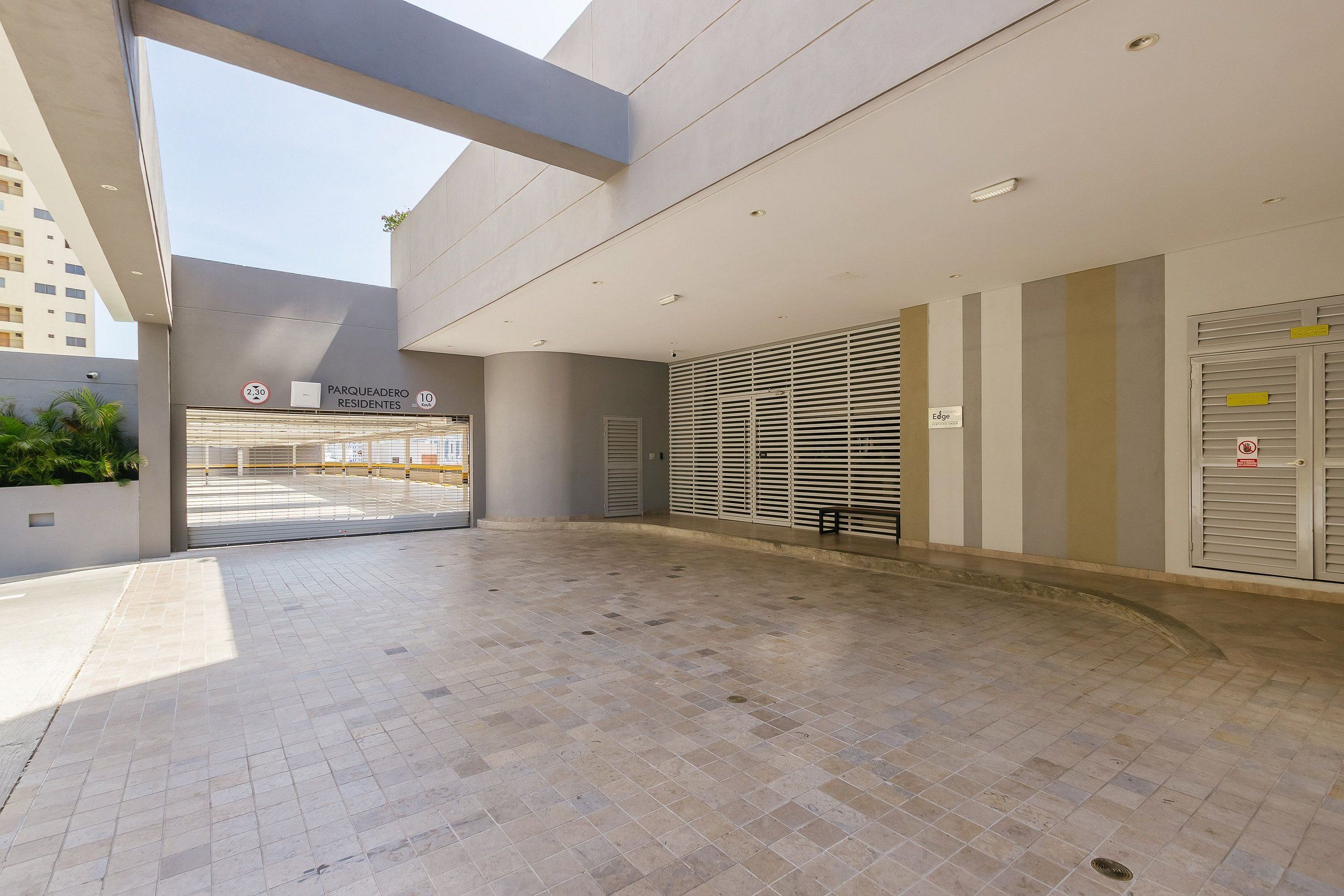 Une entrée d’immeuble spacieuse et moderne, avec un sol carrelé, un panneau indiquant une zone de stationnement réservée aux résidents et un espace d’attente abrité équipé de stores.