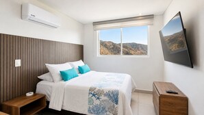 A bedroom with a bed, a bedside table, a nightstand, a wall-mounted TV, and a window with a view of hills. - U-GO Edificio Ambar Roca en Rodadero Sur (Santa Marta)
