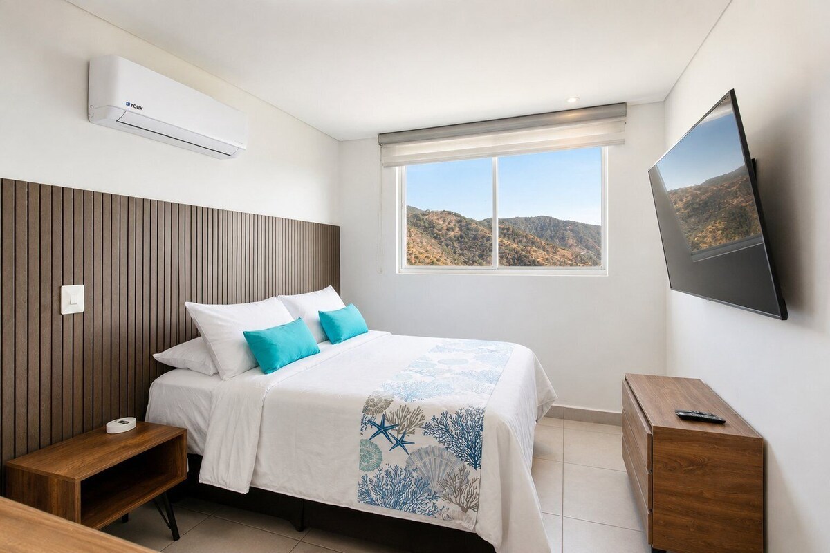 A bedroom with a bed, a bedside table, a nightstand, a wall-mounted TV, and a window with a view of hills.