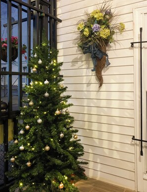 A Christmas tree decorated with ornaments, positioned next to a white wall with a hanging flower arrangement.