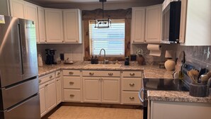 A modern kitchen with white cabinets, a stainless steel refrigerator, a built-in oven, a sink, and a window with blinds. - Sturgis Cozy Cottage (Sturgis)