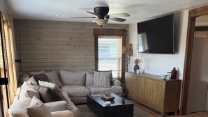 A modern living room with a sectional sofa, ottomans, a coffee table, a ceiling fan, a flat-screen TV, and a wooden cabinet. - Sturgis Cozy Cottage (Sturgis)