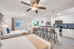 A modern hotel room with a large bed, a desk with chairs, a kitchenette, and a ceiling fan.