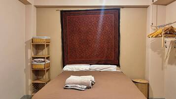 Un lit bien fait, recouvert d’une couverture marron et agrémenté d’une serviette blanche pliée, une étagère en bois avec des paniers, et une grande tapisserie à motifs accrochée au-dessus du lit.