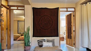 Un cuarto amplio con una cama grande, una mesita con objetos decorativos, una planta en maceta y un tapiz colgado en la pared.
