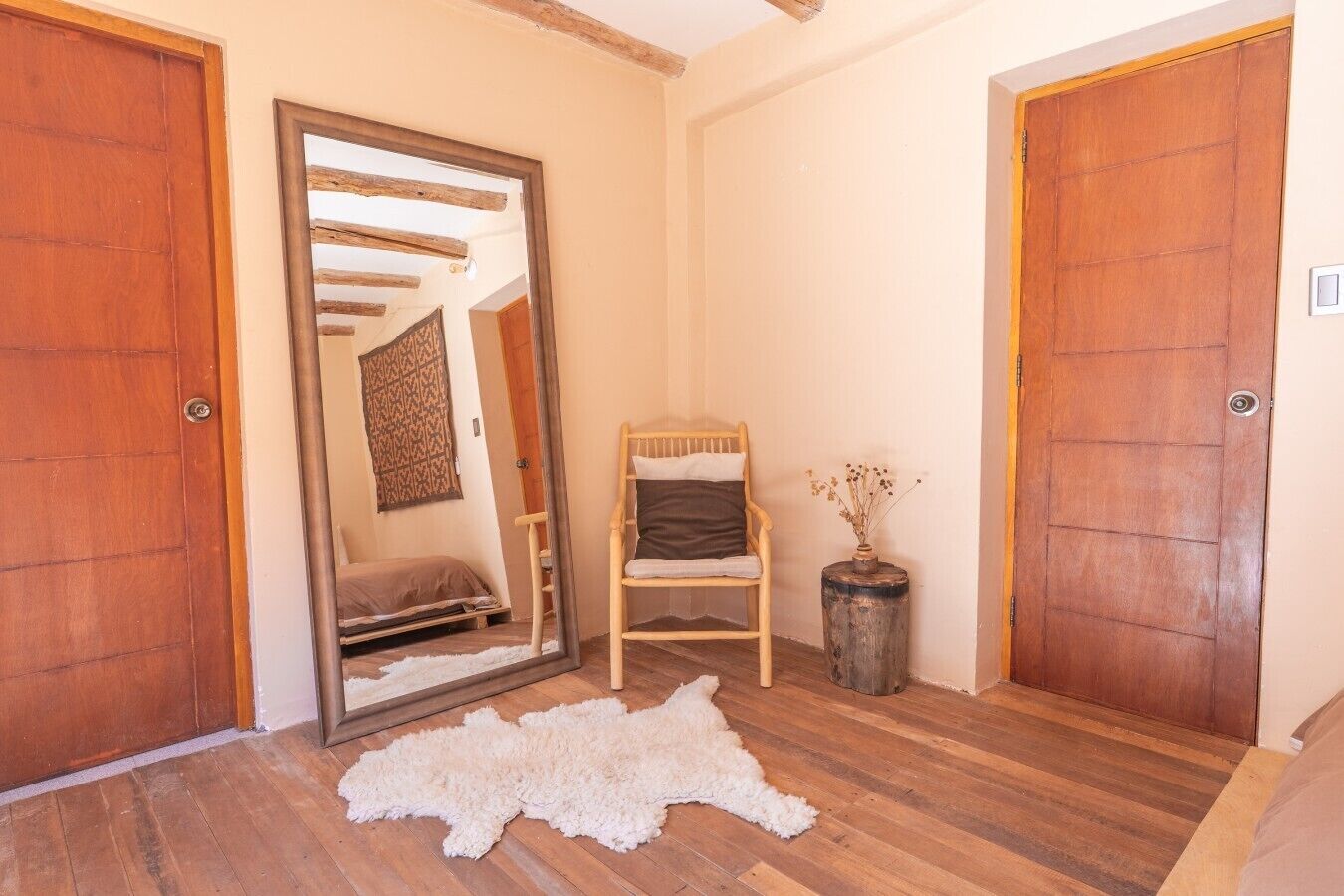 A room with wooden flooring, a white fur rug, a wooden chair, a small table with a vase, and two wooden doors.