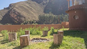A rustic outdoor area with a fire pit surrounded by wooden stumps, a mountain backdrop, and a building with a circular window.