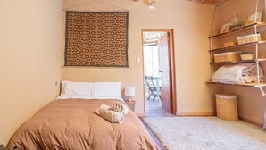A bedroom with a bed, a wall hanging, a shelf with towels, and a door leading to another room.