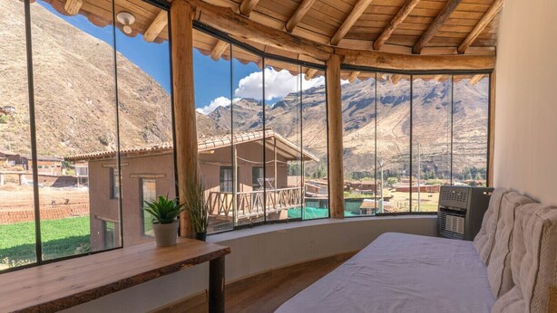 A room with a bed, a wooden bench, and a large window offering a mountain view.